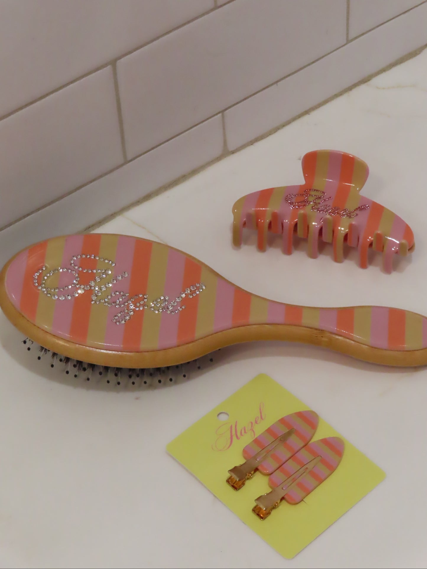 Striped pink and orange hairbrush, claw clip, and matching hair clips with sparkling cursive lettering on a white countertop.