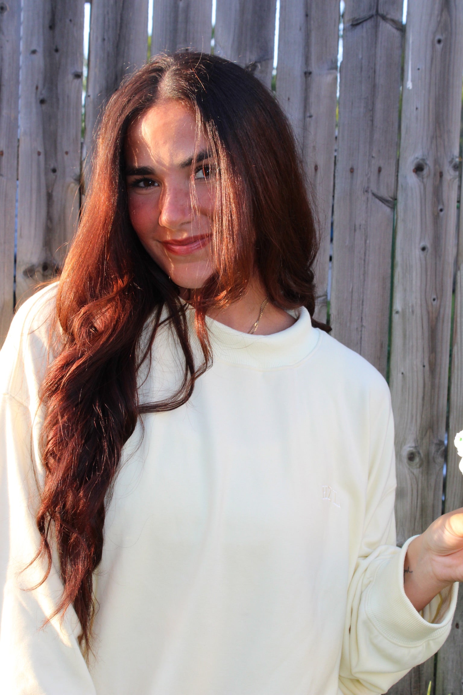 Young woman with long brown hair wearing a soft banana-colored mockneck top standing in front of a wooden fence smiling casually