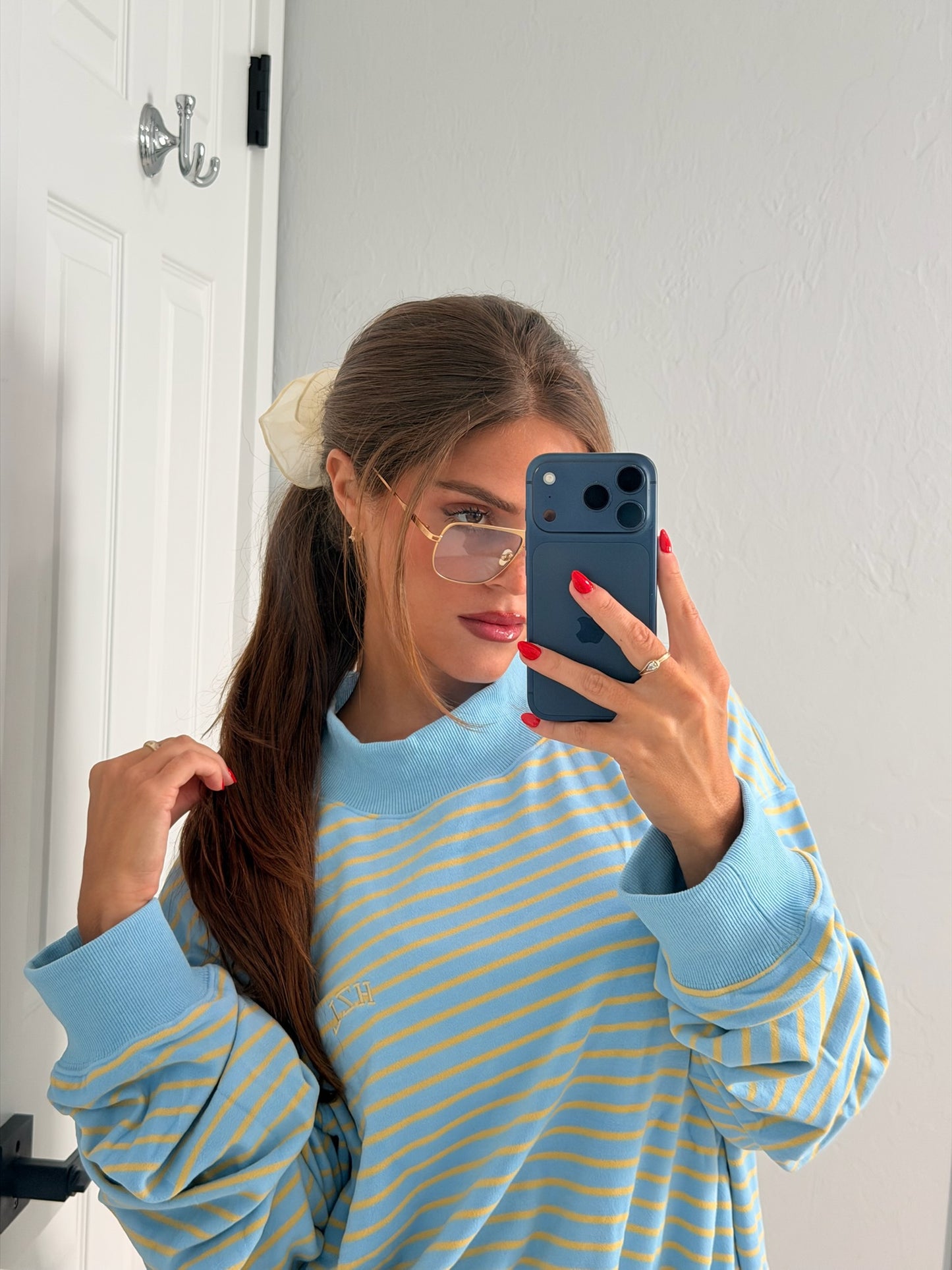 Young woman with long brown hair tied with a cream scrunchie, wearing gold-rimmed glasses and a blue and yellow striped mockneck sweater taking a mirror selfie with a dark blue smartphone.
