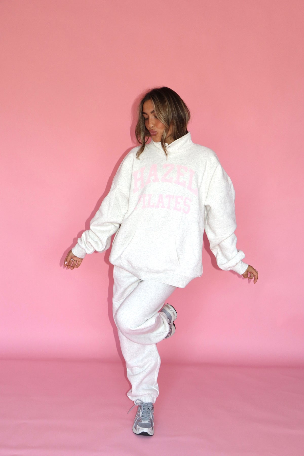 Woman wearing white Pilates joggers and a matching hoodie against a pink studio backdrop