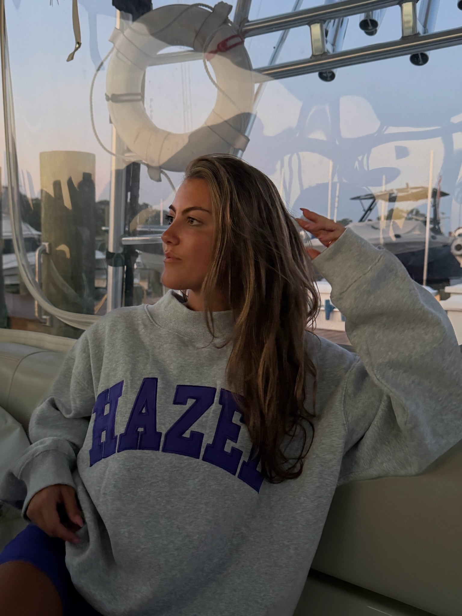 Woman wearing a cozy gray sweatshirt with bold purple lettering, relaxing on a boat at sunset.