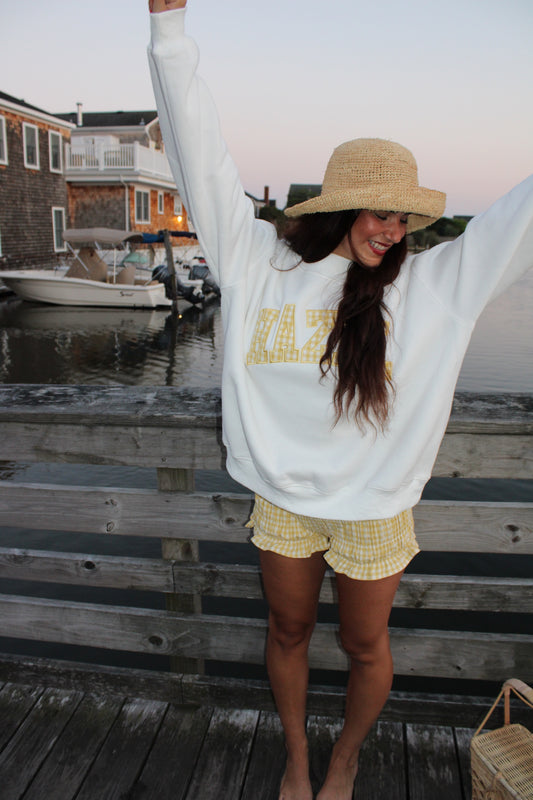 Young woman wearing yellow gingham shorts and a white sweatshirt with a straw hat standing on a dock by the water at sunset.