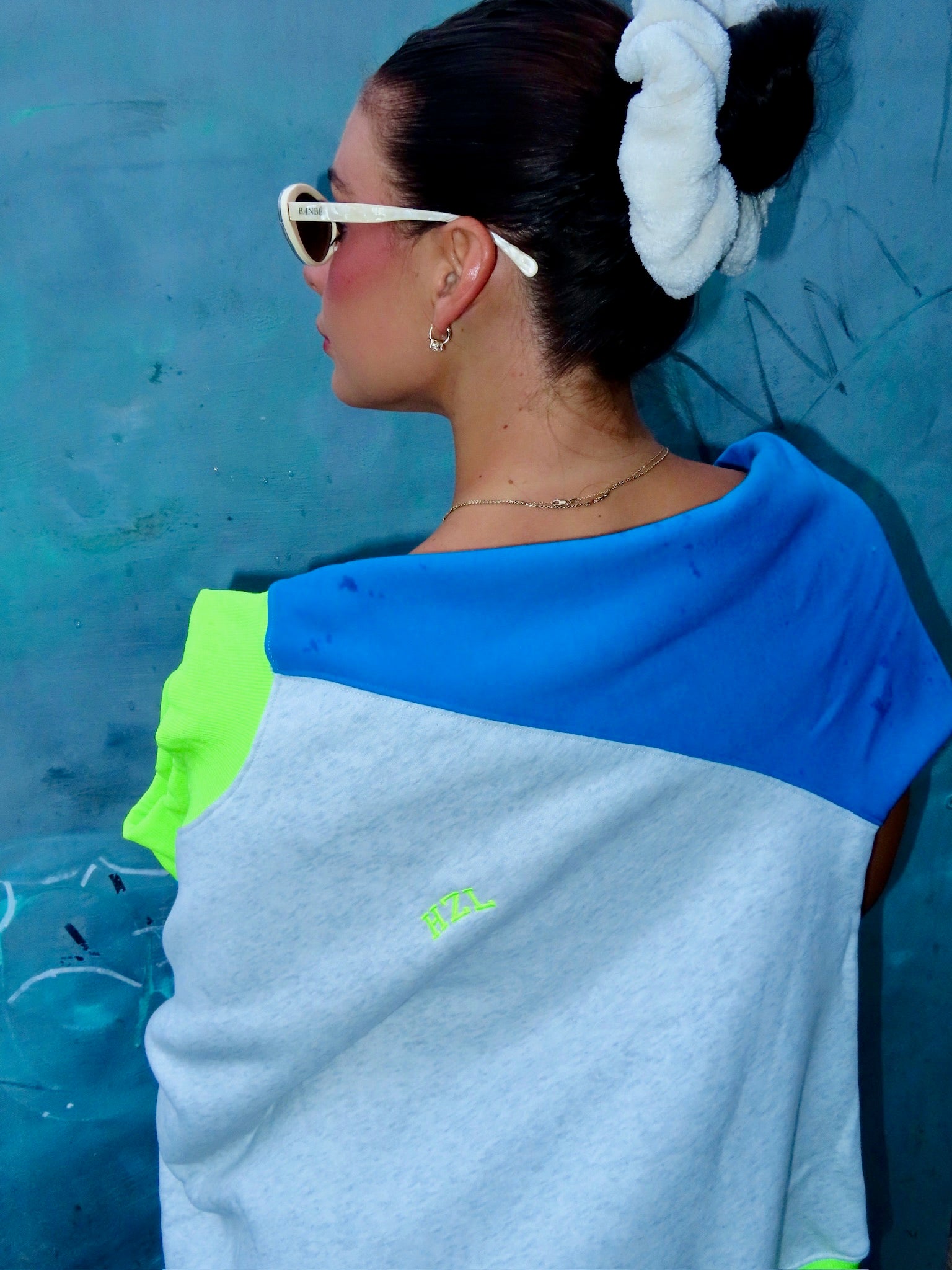 Woman wearing a colorful mockneck top with blue, gray, and neon green panels, styled with white hair scrunchie and sunglasses