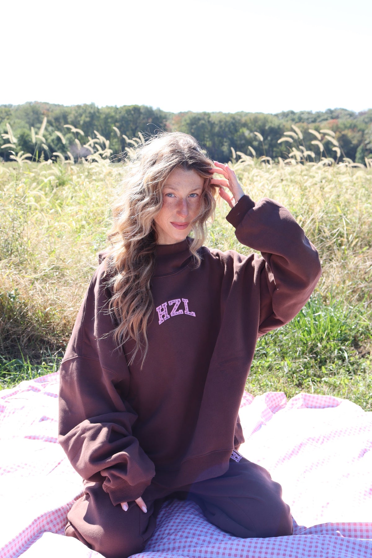 Woman wearing a dark brown HZL mock hoodie seated on a picnic blanket in a sunny field
