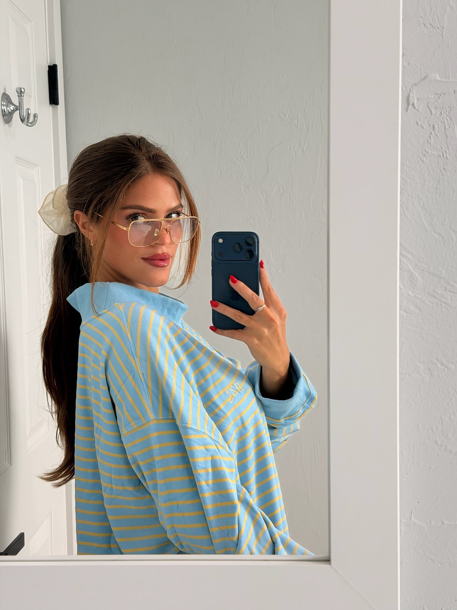 Young woman with long hair tied back with a scrunchie, wearing oversized striped top and clear glasses, taking a mirror selfie with a phone.