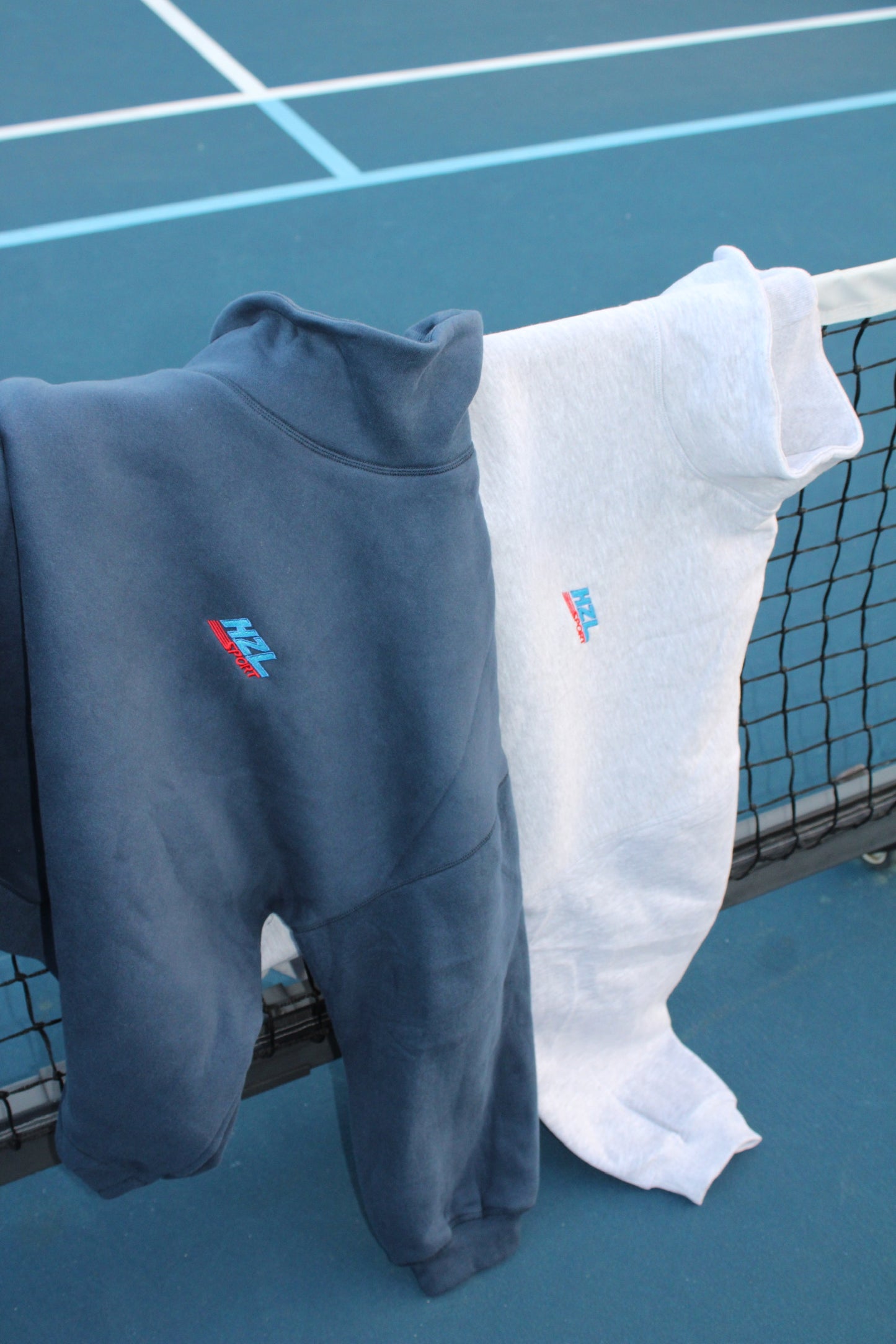 Navy and white sporty mockneck sweatshirts hanging over a tennis net on an outdoor court.