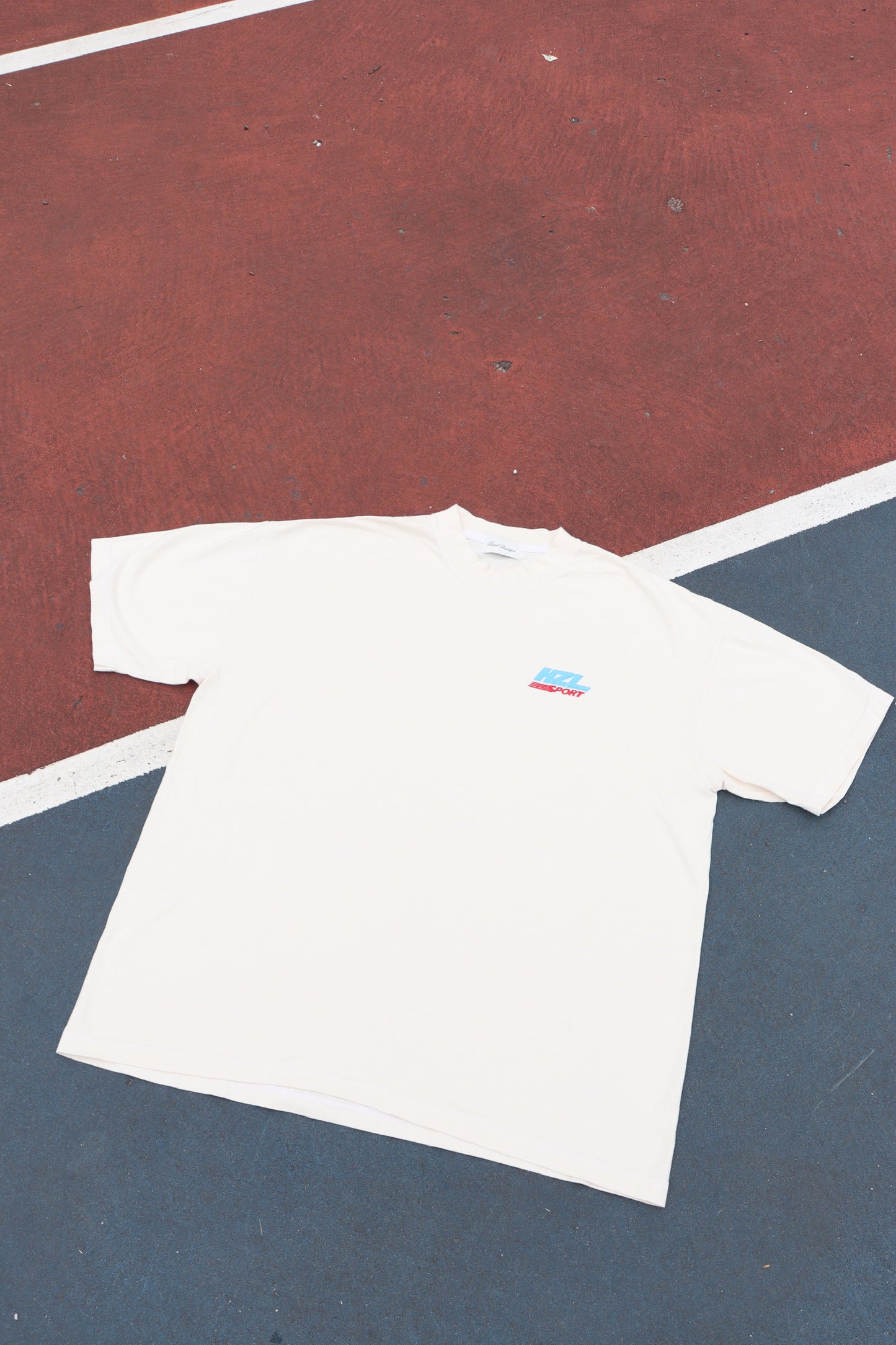 Cream short sleeve tee laid flat on a red and blue court with a small colorful chest graphic
