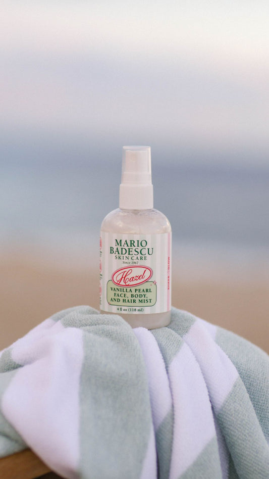 Clear spray bottle of vanilla pearl mist for face, body, and hair sitting on a striped towel outdoors at the beach.