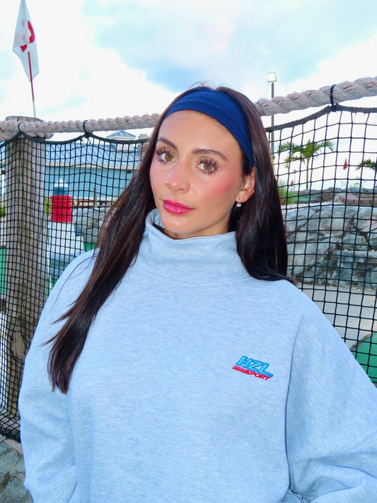Woman wearing a light grey sporty mockneck sweatshirt with a blue headband, standing outdoors by a net fence.