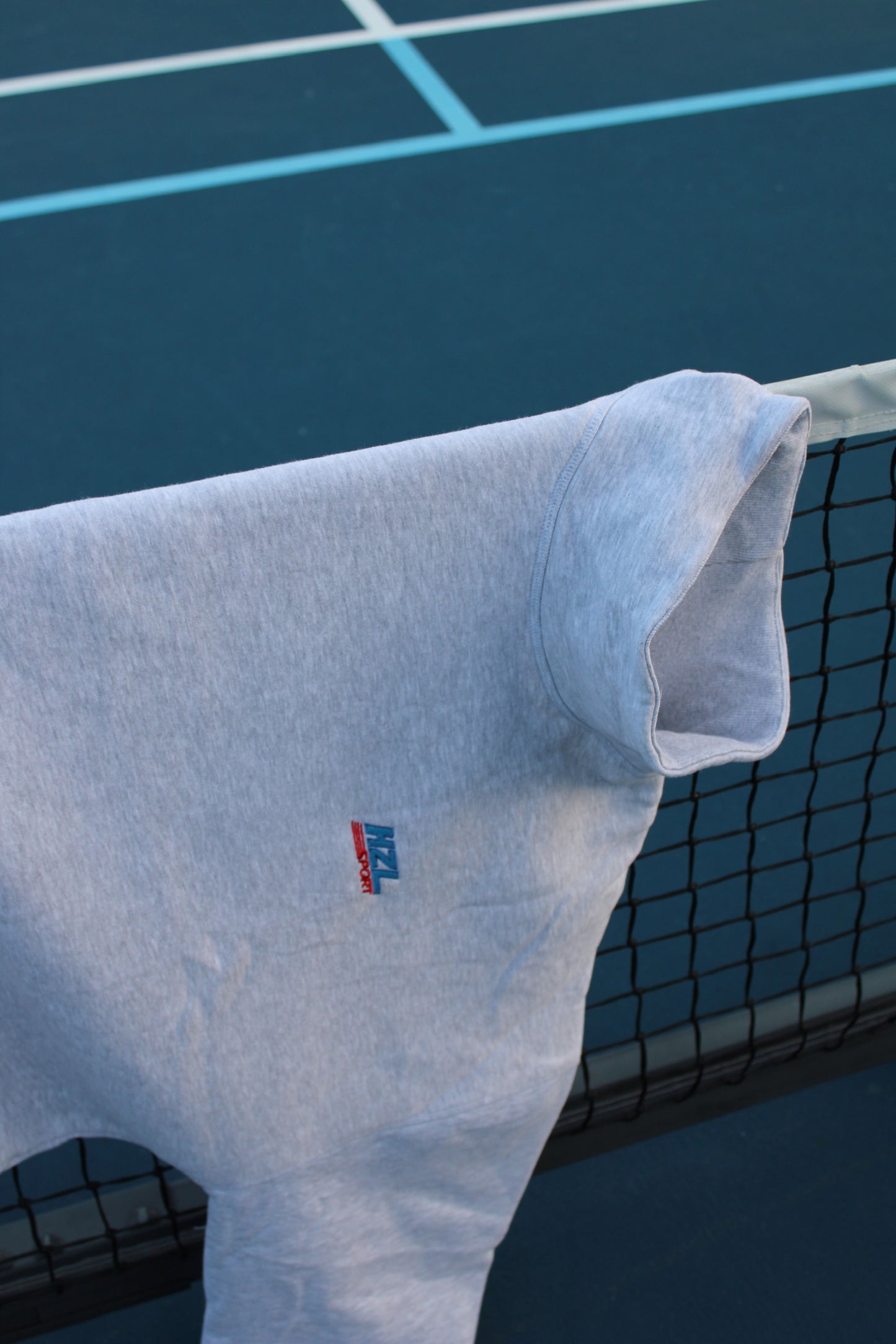 Grey sporty mockneck with small logo detail draped over a tennis net on a blue court