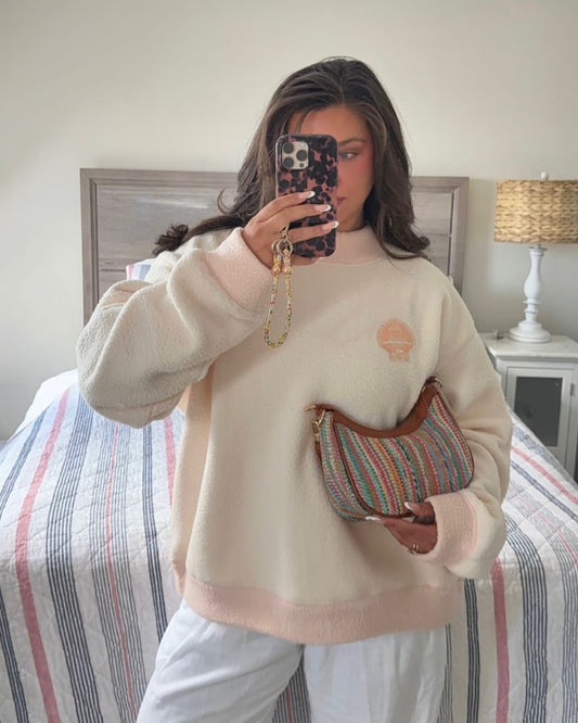 Woman wearing a cozy cream and blush fleece pullover holding a striped woven handbag in a neutral bedroom setting