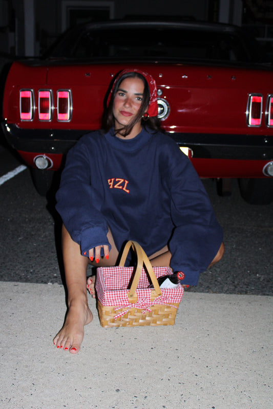 Young woman wearing a navy blue HZL sweatshirt and red bandana sits barefoot on pavement next to a picnic basket in front of a red vintage car at night.