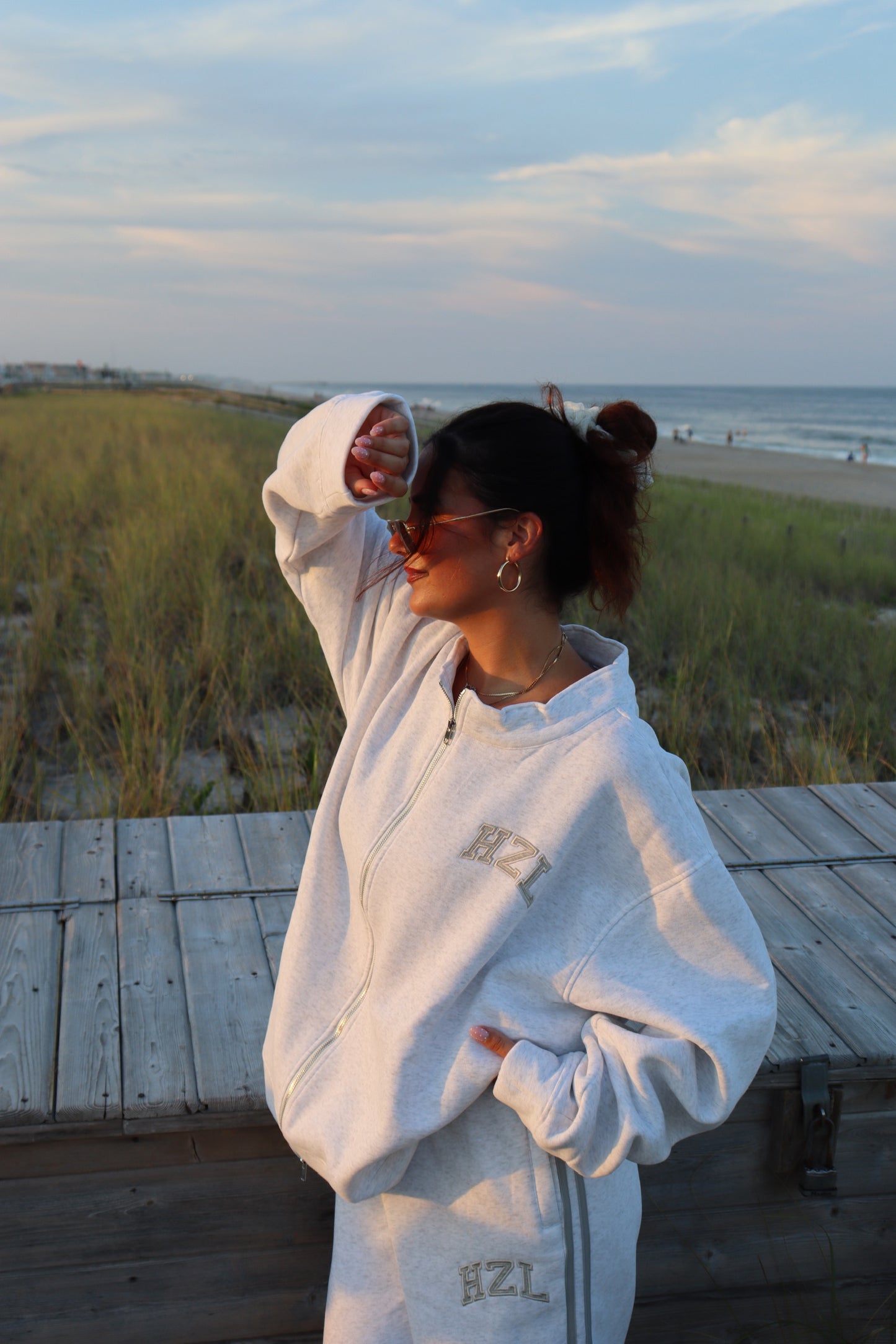 Woman in a light gray HZL zip-up sweatshirt standing on a beach boardwalk beside the ocean