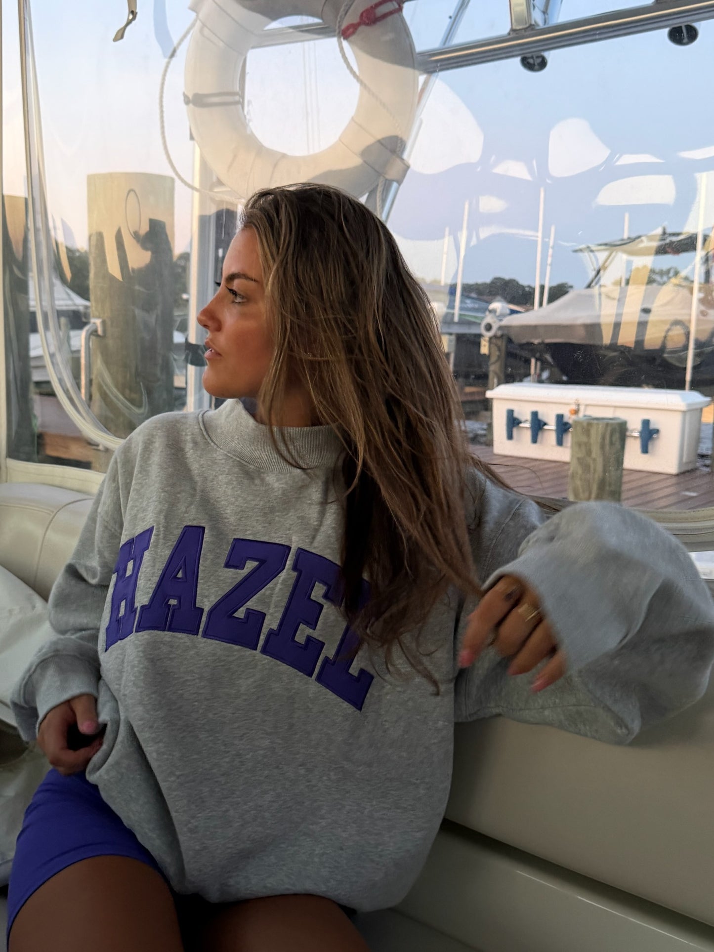 Woman with long hair wearing a gray sweatshirt with purple lettering sitting on a boat near the dock
