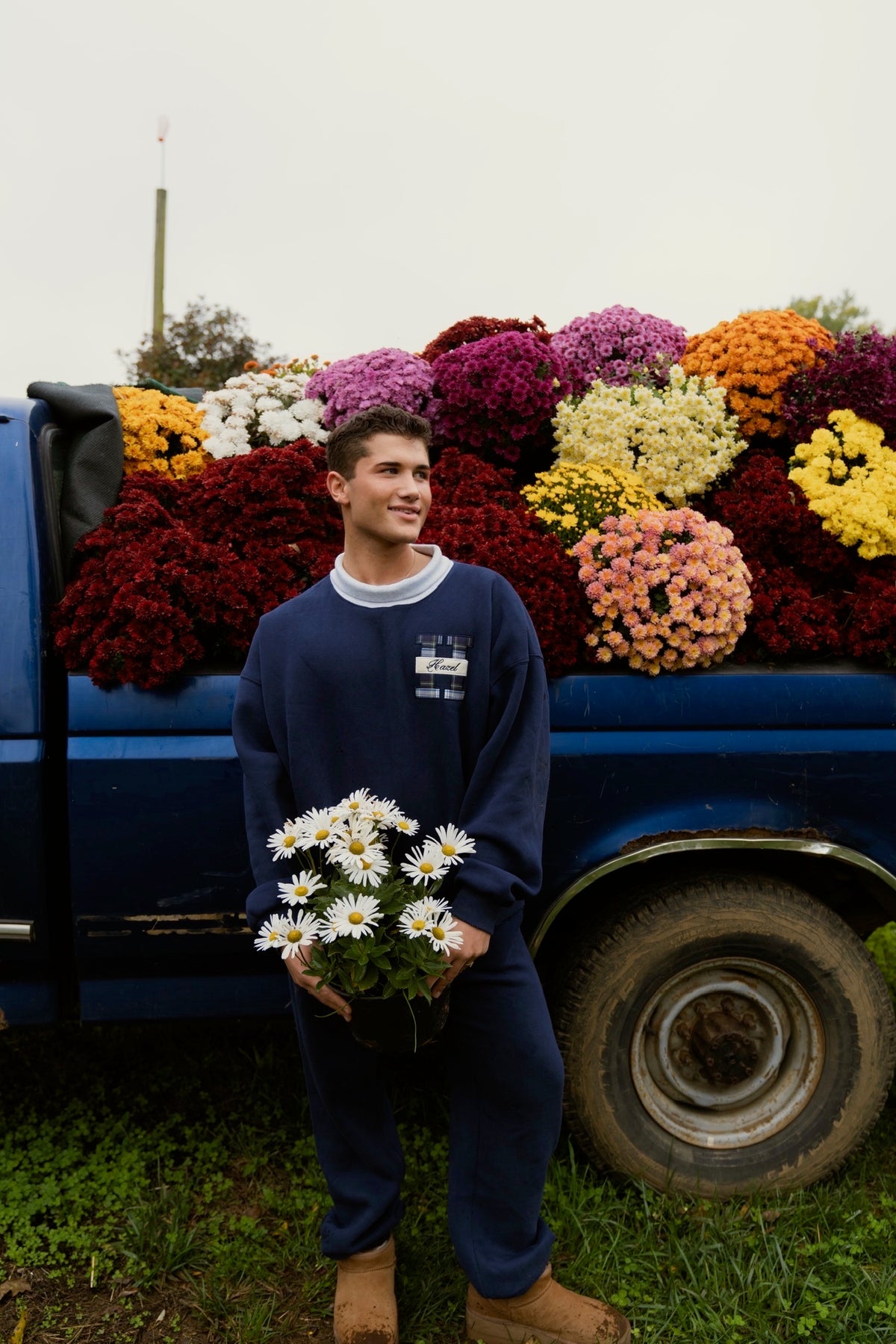 Man wearing a navy mockneck sweatshirt and matching pants holds daisies beside a colorful flower truck