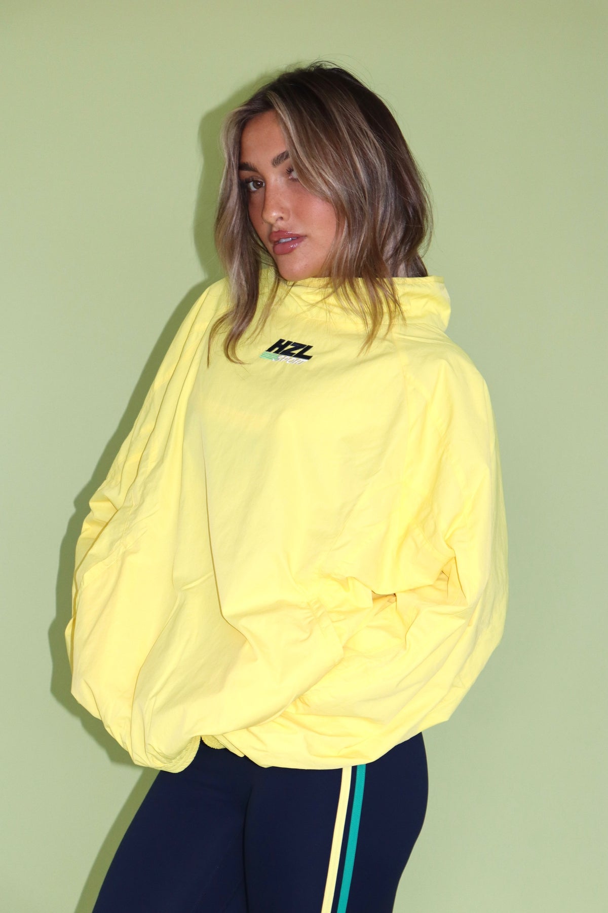 Woman wearing a yellow HZL windbreaker with navy athletic leggings against a green background