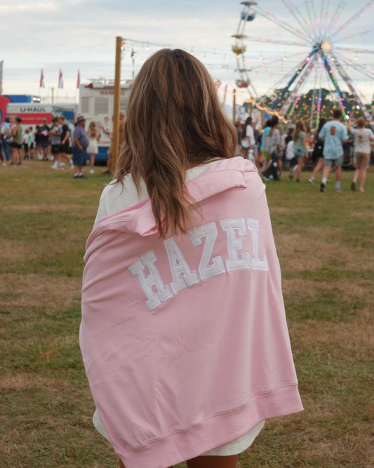 Woman draped in a pink sweatshirt with "HAZEL" on the back at a lively outdoor fair with a ferris wheel in the background