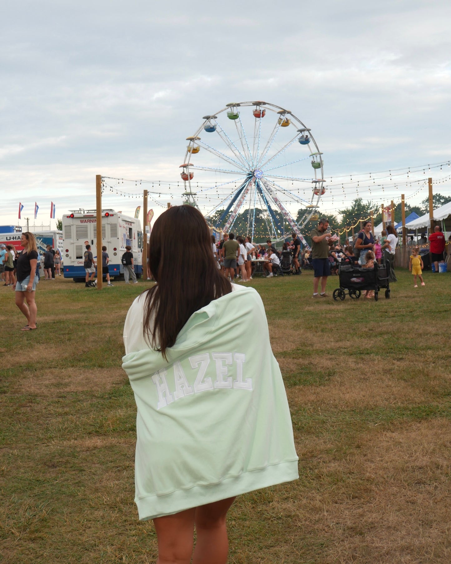 Woman wrapped in a mint green Hazel Boutique sweatshirt with a Ferris wheel and fair crowd in the background