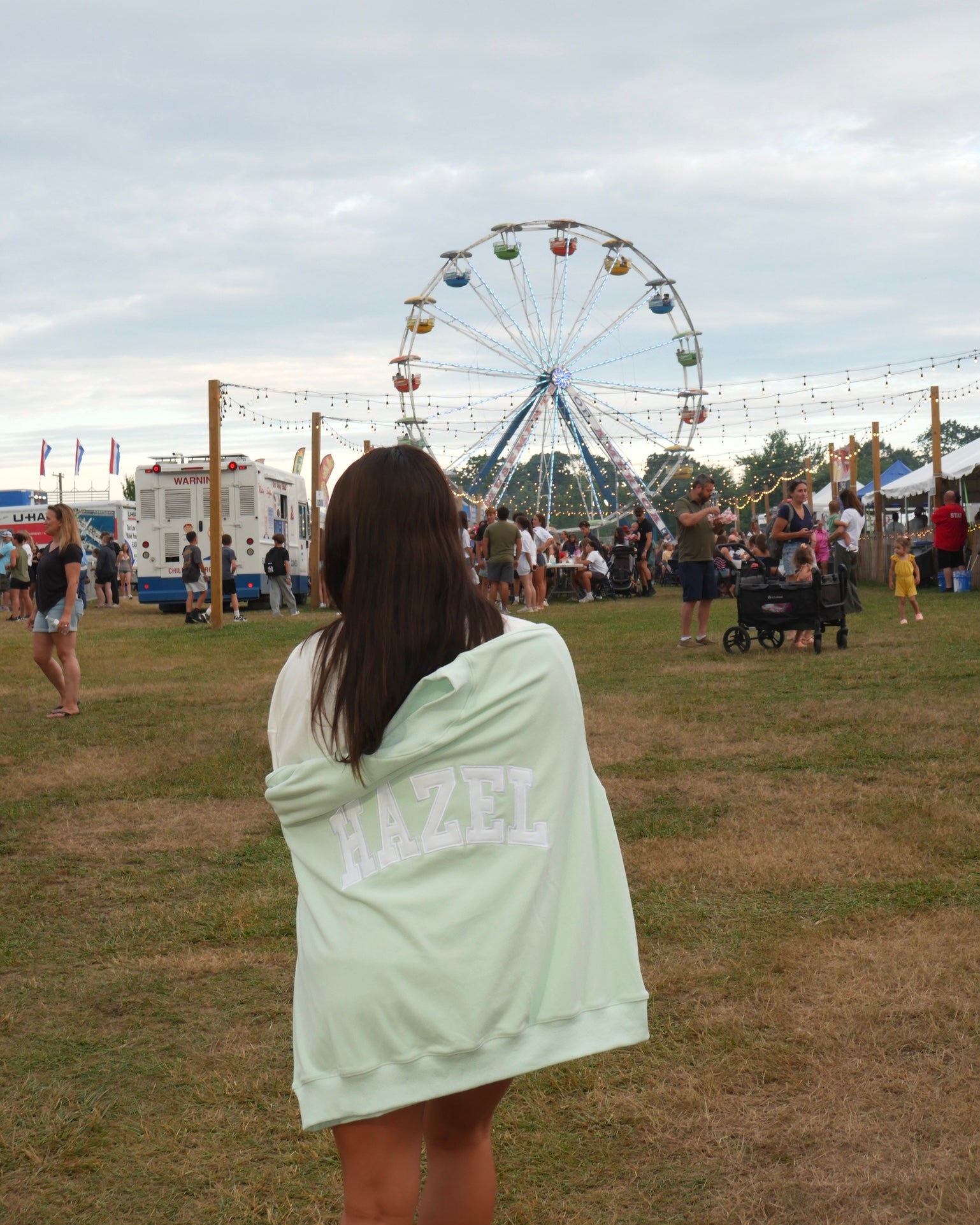 Woman wrapped in a mint green Hazel Boutique sweatshirt with a Ferris wheel and fair crowd in the background