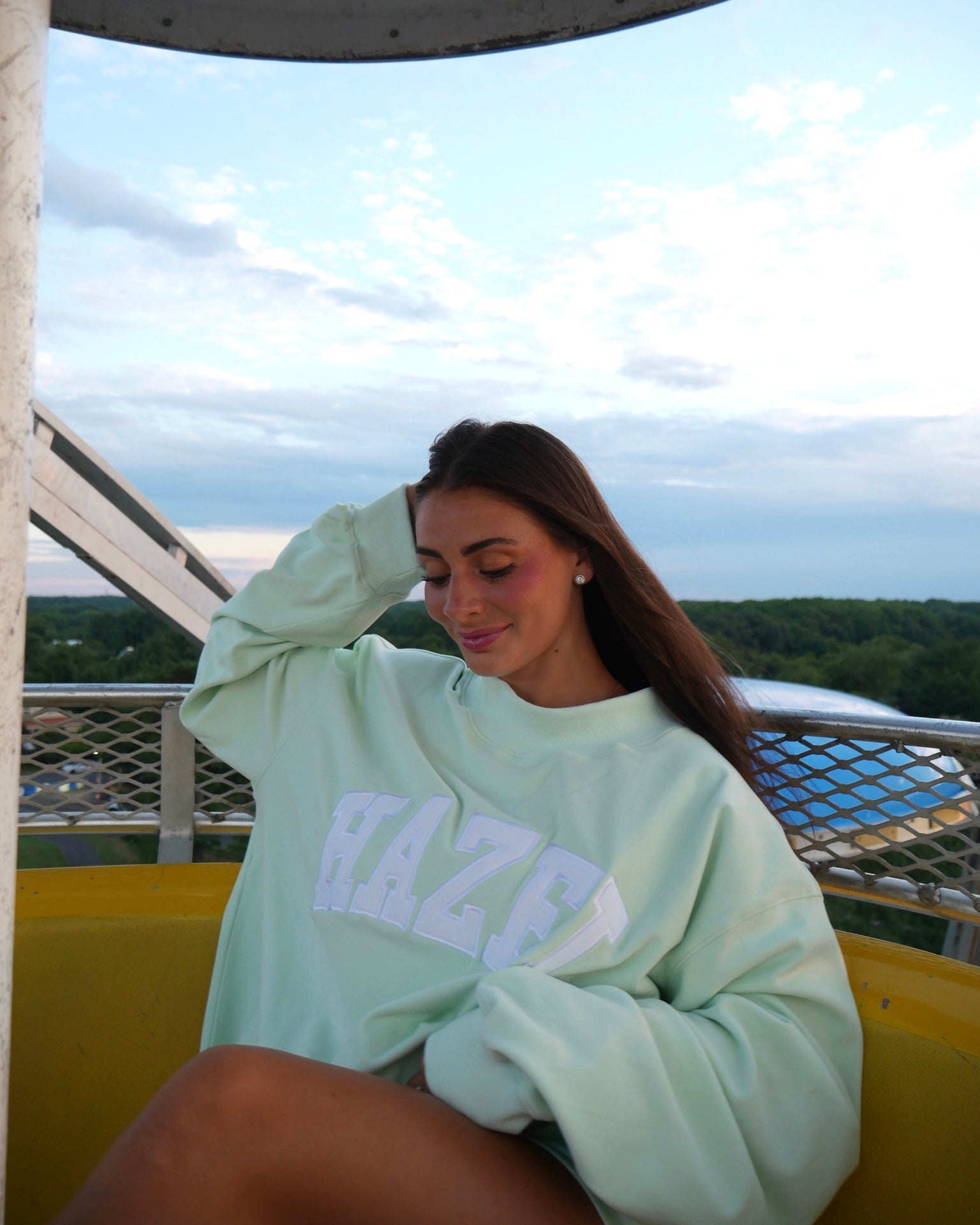 Young woman wearing a mint green oversized sweatshirt with lettering, sitting outdoors on a Ferris wheel at sunset.