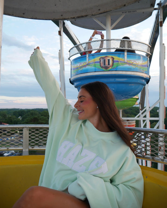 Young woman smiling with arm raised wearing a mint green mockneck sweater on a carnival ride during sunset