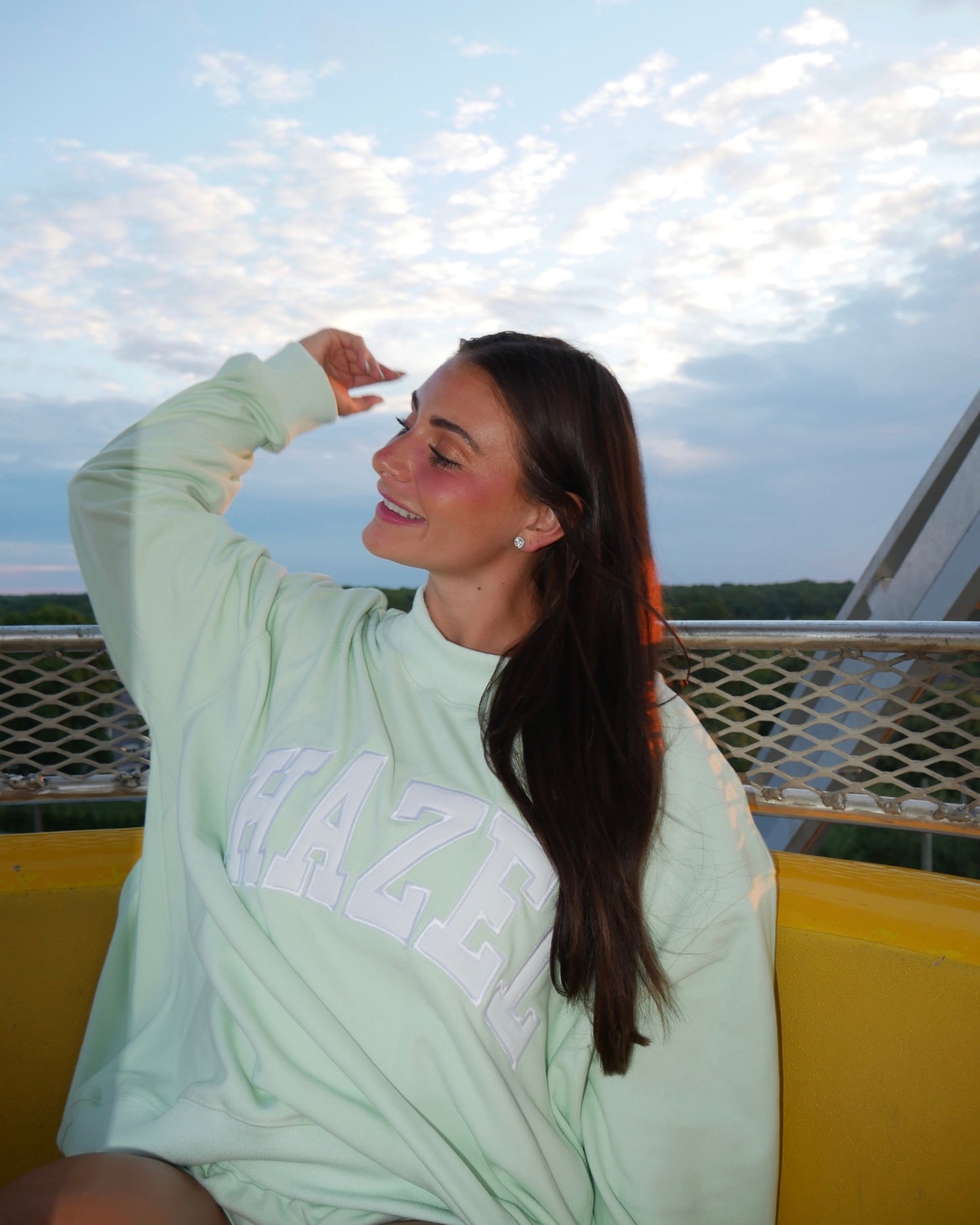 Young woman wearing a mint green oversized sweatshirt with white lettering, smiling outdoors at sunset.