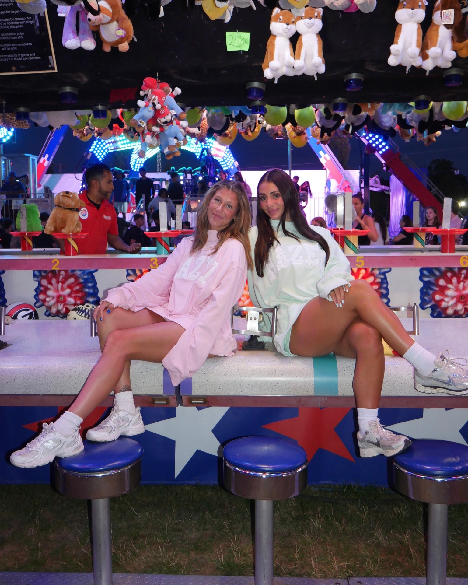 Two young women wearing casual long-sleeve tops sitting on stools at a colorful carnival game booth at night.