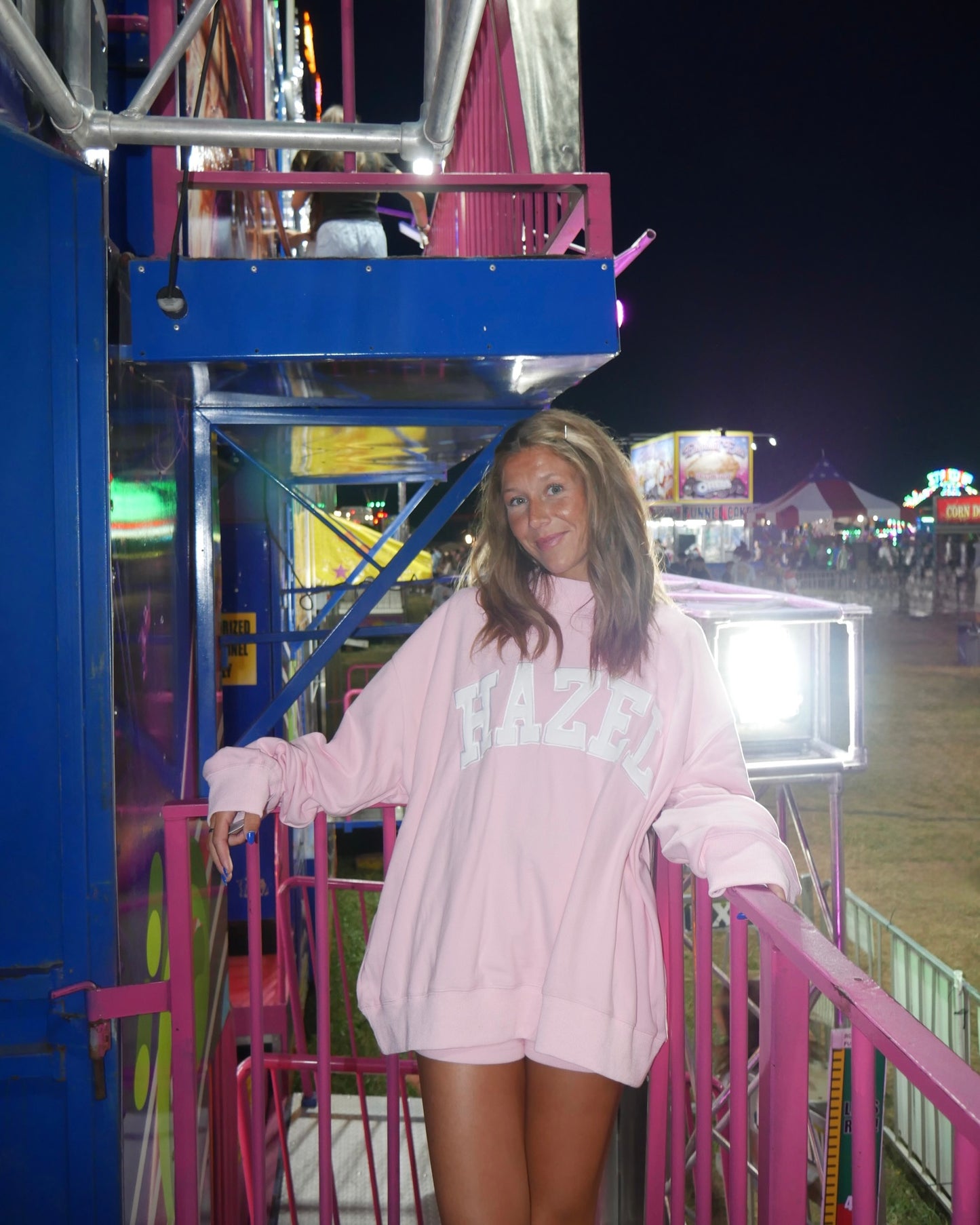 Woman wearing a pink oversized mockneck sweatshirt with Hazel lettering standing on a colorful carnival ride at night